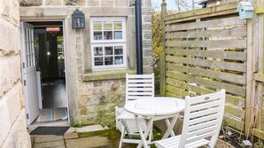 A stone building with a white door, a window, and a small outdoor seating area with a table and chairs. - Ash Cottage (Bakewell)