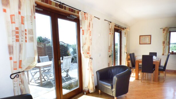 Il s’agit d’une pièce spacieuse dotée d’une grande porte-fenêtre coulissante qui donne sur un patio extérieur. À l’intérieur, on trouve deux fauteuils en cuir noir et une table à manger avec des chaises.