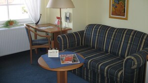 Una habitación de hotel con un sofá a rayas, una mesita redonda con una revista, una silla, una lámpara y una ventana con cortinas.