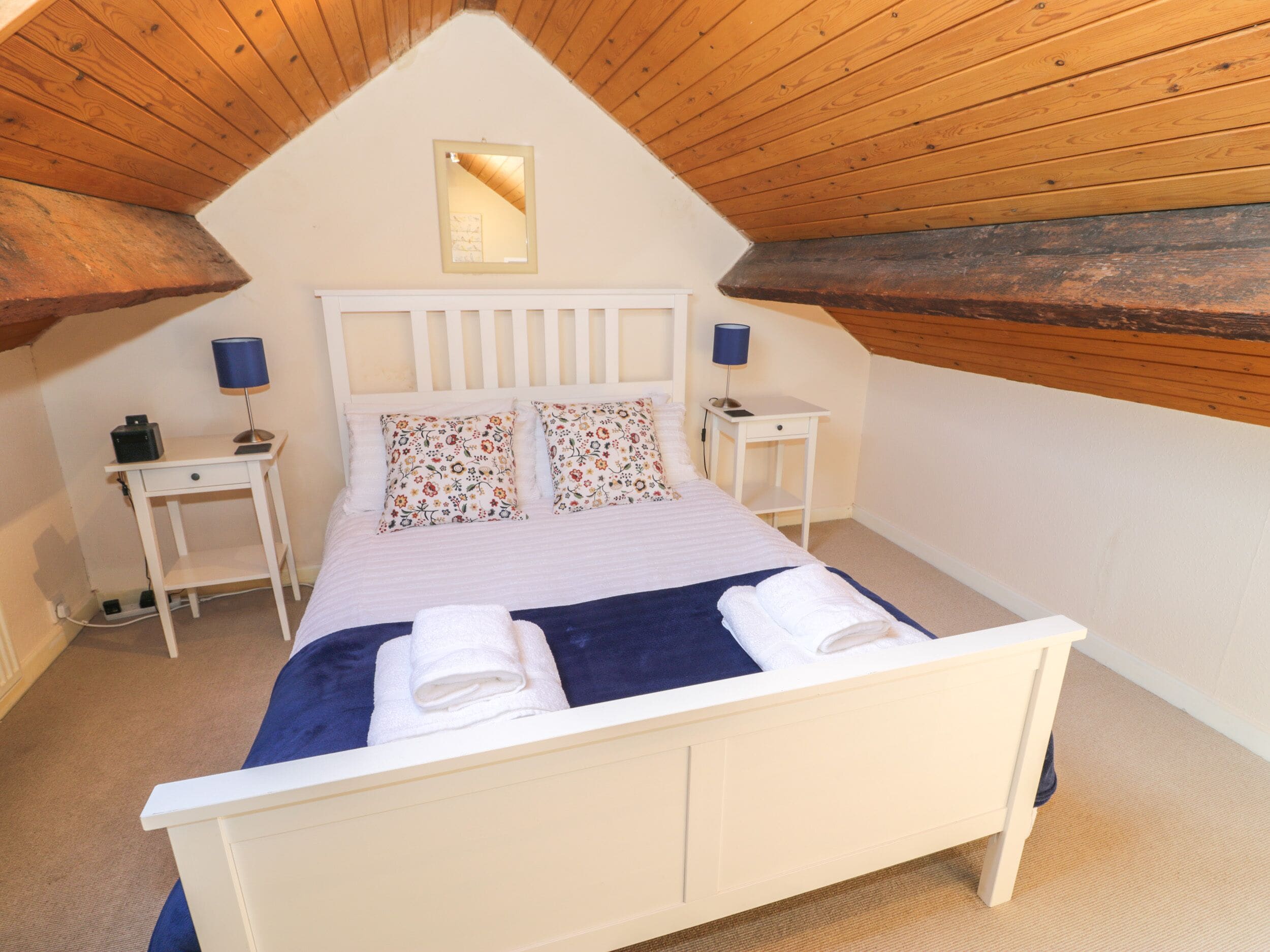 A bedroom with a sloped ceiling, a bed with floral pillows, two bedside tables with lamps, and a small mirror on the wall.
