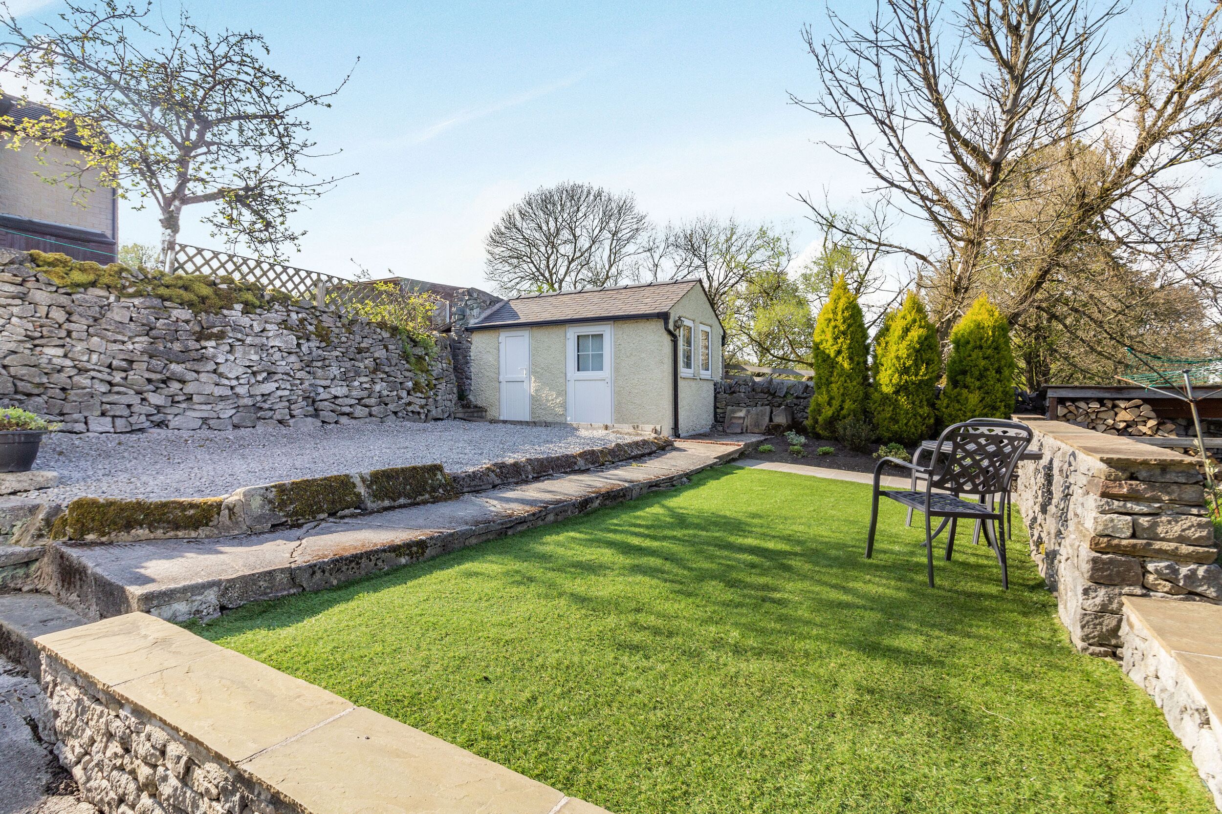 Un jardin entouré de murs en pierre, avec une petite maison, une pelouse et un espace aménagé pour s’asseoir.