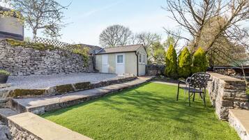 Un jardin entouré de murs en pierre, avec une petite maison, une pelouse et un espace aménagé pour s’asseoir.