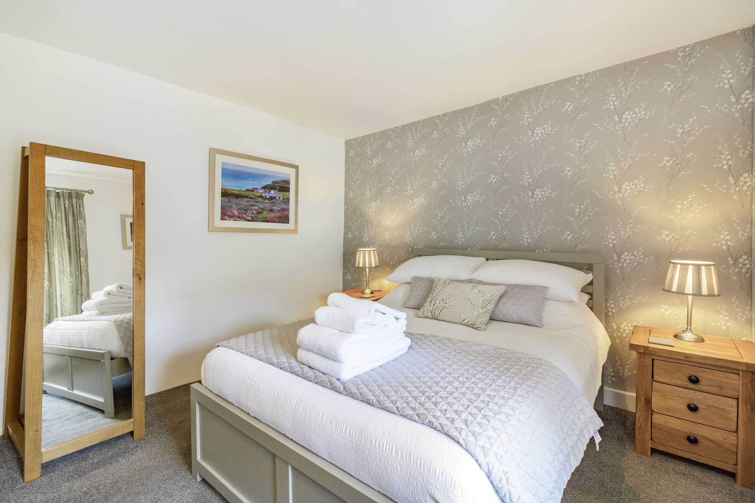A neatly made bed with white linens, a bedside table with a lamp, and a wall with a floral pattern.