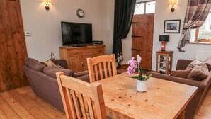 Un salon chaleureux avec une table à manger en bois, deux chaises en bois, un canapé et un téléviseur posé sur un meuble en bois.