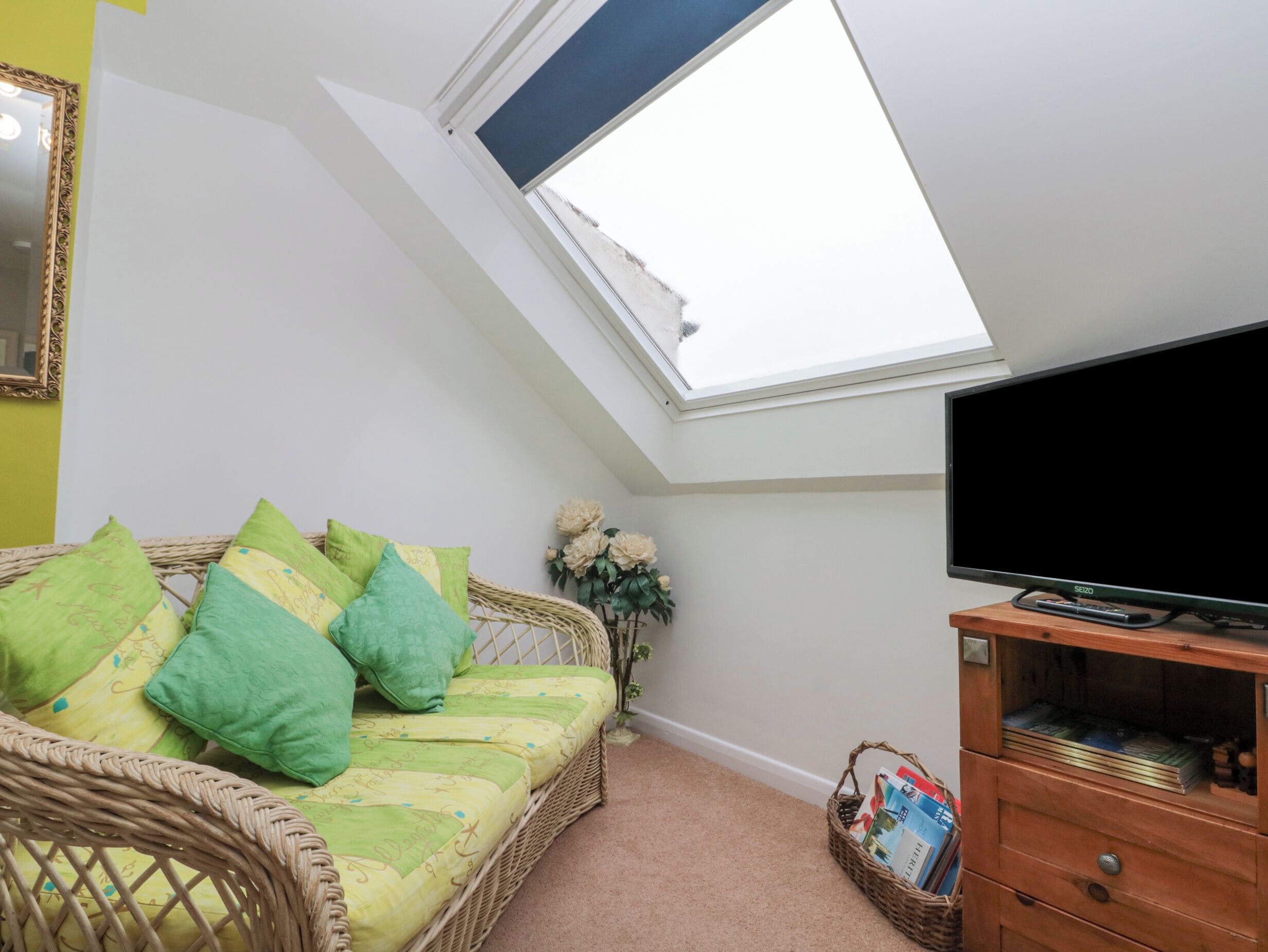 A room with a sloped ceiling, a television on a wooden stand, a wicker sofa with green cushions, and a small side table with a basket.