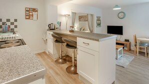 Una cocina moderna con una isla central, taburetes de bar, un área de comedor con televisor y piso de madera.