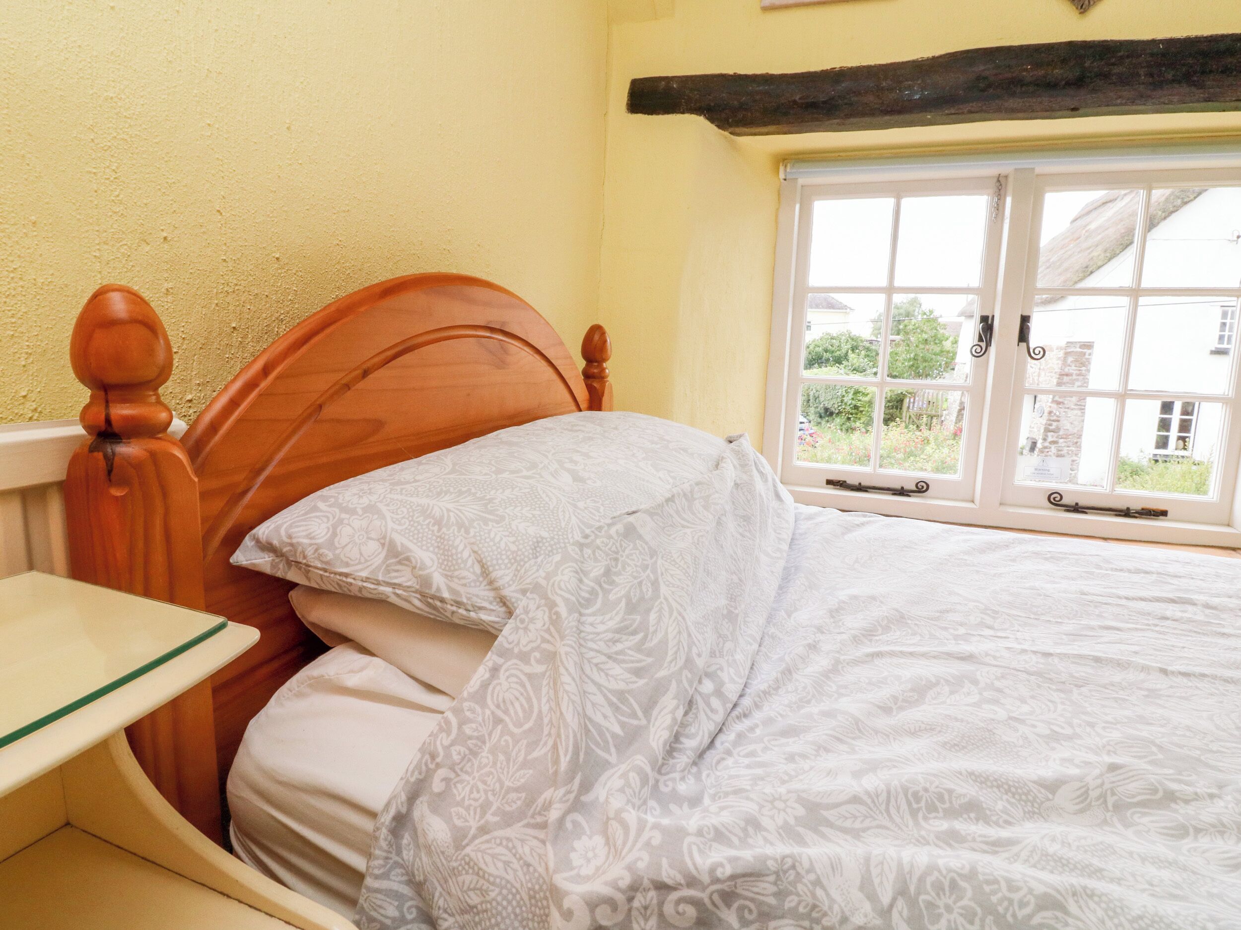 A bed with a wooden headboard, a bedside table, and a window with a view of buildings and greenery.