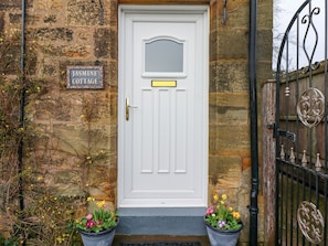 A white door with a small window above it, flanked by two flower pots on either side. - Jasmine Cottage (Alnwick)