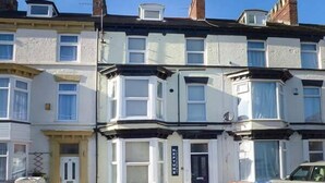 A row of terraced houses with bay windows, a front door, and a small garden area with a parked car and bins. - Flat 1 (Bridlington)