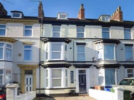 A row of terraced houses with bay windows, a front door, and a small garden area with a parked car and bins.