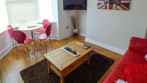 A living room with a red sofa, a wooden coffee table, a TV mounted on the wall, and a painting of a Union Jack. - Flat 1 (Bridlington)