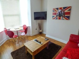 Un salón con un sofá rojo, una mesa de centro de madera, un televisor montado en la pared y un cuadro con la bandera del Reino Unido.