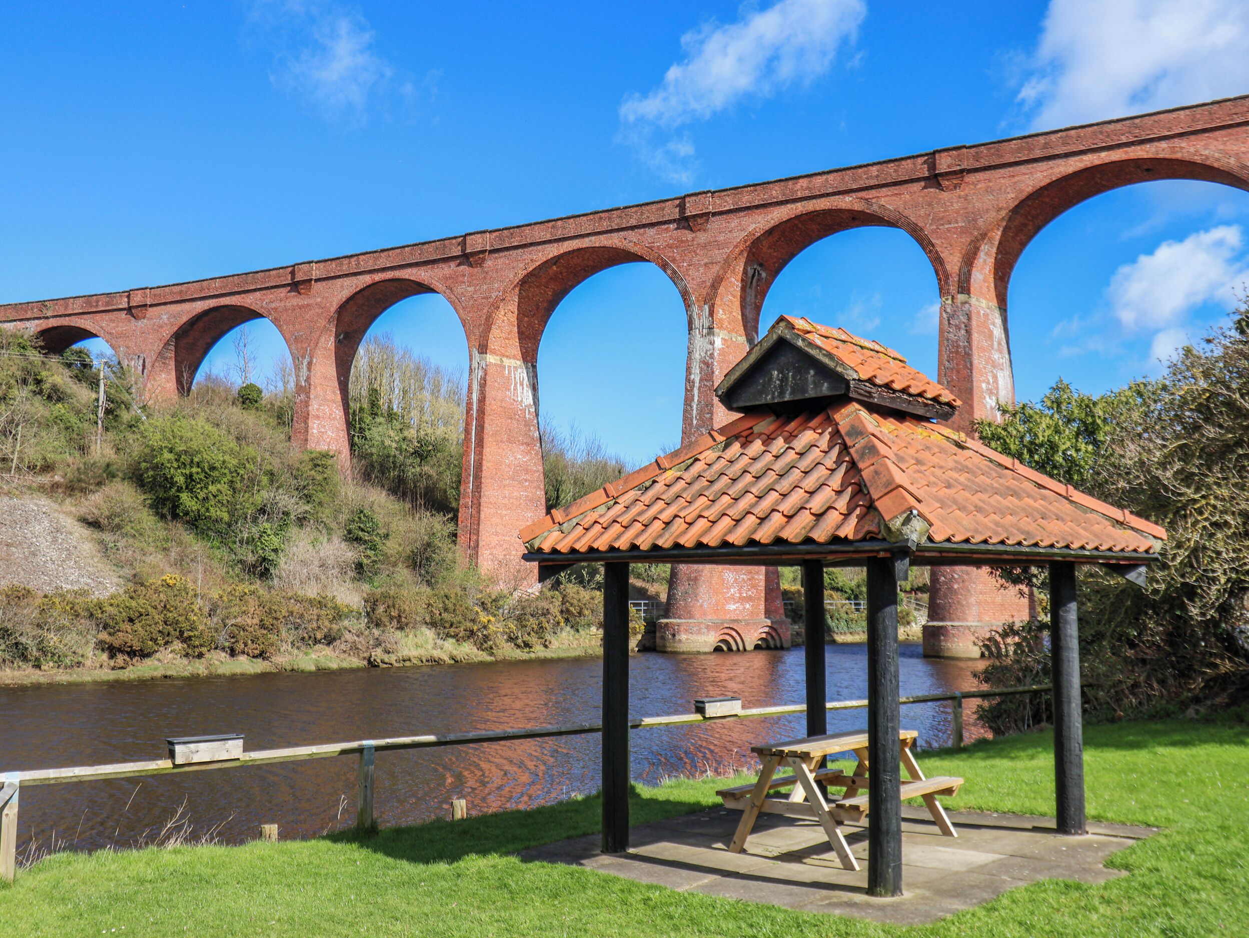 Un área de picnic con bancos y un pabellón, un río y un puente de ladrillo con arcos.