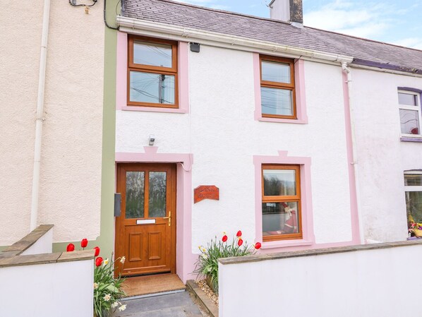Une maison à deux étages, avec une façade blanche et des ornements roses, une porte d’entrée en bois et un petit jardin où poussent des tulipes rouges.