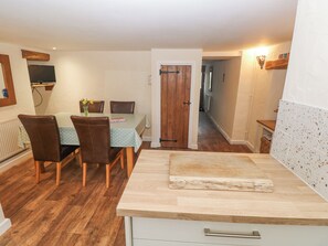 Un coin repas avec une table et des chaises en bois, un comptoir de cuisine avec une planche à découper, et un couloir menant à une autre pièce.