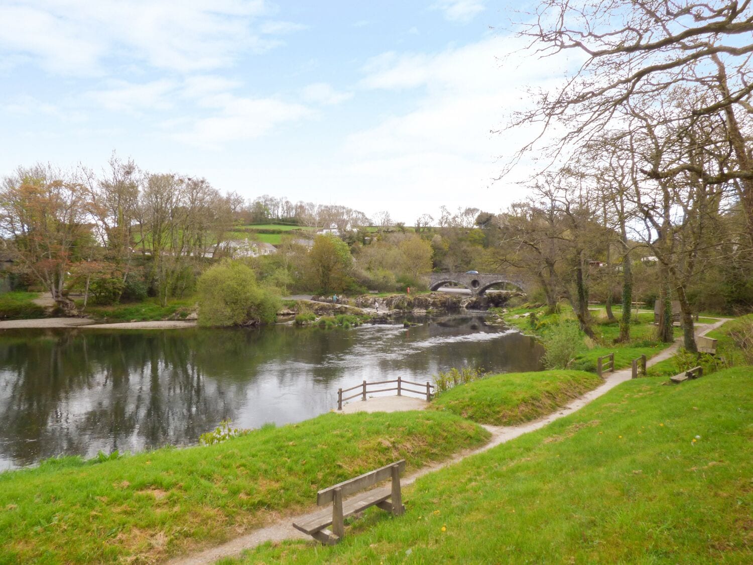 A riverside park with a wooden bridge, a bench, and a path.