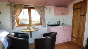 A cozy room with a bed, two chairs, a small table with wine glasses, a window with curtains, a kitchenette with a sink, and a wooden door. - Llety'r Bugail (Maesteg)