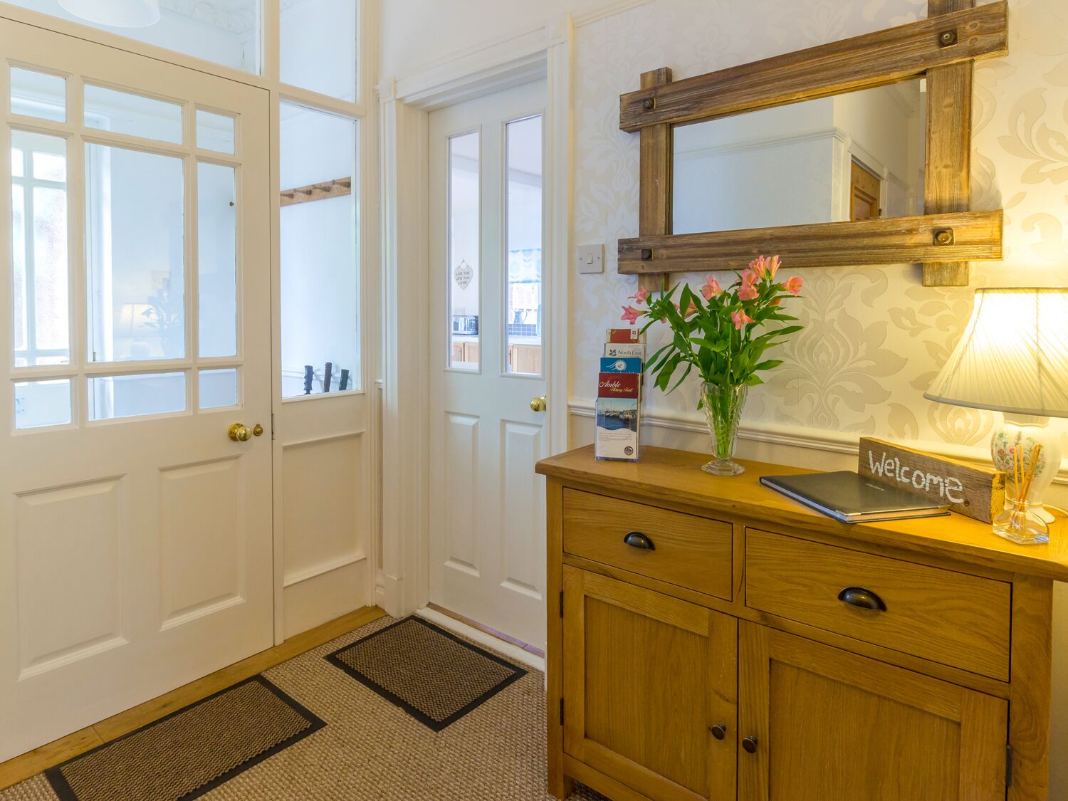 A hotel reception area with a wooden cabinet, a welcome sign, a vase with flowers, a lamp, and a mirror.