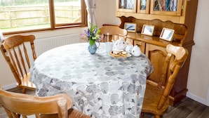 A dining area with a round table covered by a tablecloth with a pattern of mushrooms. There are chairs around the table, a cabinet with glass doors, and a window with a view of a fence outside. - The Dovecote (Bideford)