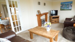 A living room with a brown leather sofa, a wooden coffee table with a vase of flowers, a television on a wooden stand, and a painting on the wall. - The Dovecote (Bideford)