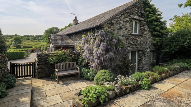 A stone house with a sloped roof, a wooden bench, and a garden with various plants and a purple flowering vine. - Kestrel Cottage (Grange-over-Sands)