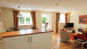 A kitchen with a wooden countertop, white cabinets, and a dining area with a chair and table. - Kestrel Cottage (Grange-over-Sands)