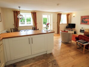 A kitchen with a wooden countertop, white cabinets, and a dining area with a chair and table. - Kestrel Cottage (Grange-over-Sands)