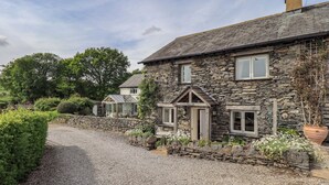 A stone house with a gravel driveway, a conservatory, and a garden with various plants. - Kestrel Cottage (Grange-over-Sands)