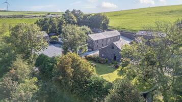 A stone farmhouse with a gravel driveway, surrounded by lush greenery and a wind turbine in the background.