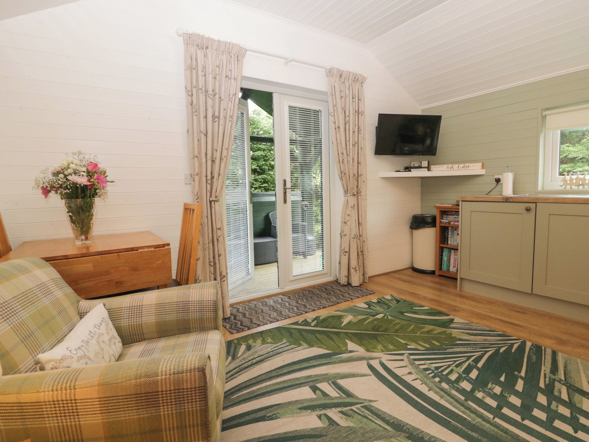 A cozy living room with a sofa, a small table with a vase of flowers, a television, and a sliding glass door leading to a balcony.
