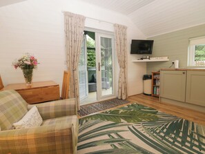 A cozy living room with a sofa, a small table with a vase of flowers, a television, and a sliding glass door leading to a balcony. - Ash Lodge (Ulverston)