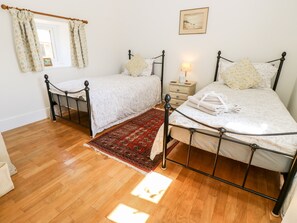 A bedroom with two single beds, a nightstand, a lamp, a window with curtains, and a rug. - The Old Dairy (Newport)