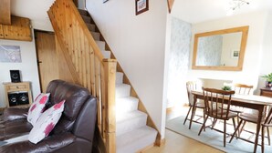 A cozy living room with a brown leather sofa, a wooden staircase, and a dining area with wooden furniture. - 60 Hyfrydle Road (Caernarfon)