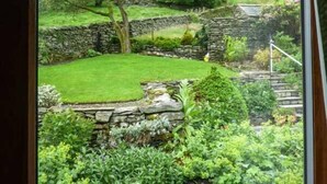 Une vue d’un jardin avec un mur en pierre, une pelouse verdoyante et diverses plantes.