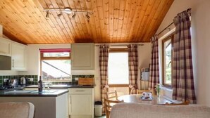 Interior acogedor de una cabaña con techo de madera, una zona de cocina con estufa y armarios, una mesa de comedor con sillas y una ventana con cortinas de cuadros.