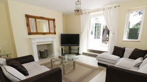 A living room with two sofas, a glass coffee table, a flat-screen TV, a fireplace, and a large window with curtains. - Garden View (Malvern)