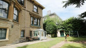 A traditional building with a conservatory, a small garden with a swing set, and a clear sky. - Garden View (Malvern)