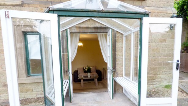 A conservatory with open doors, a sofa, a table, and a vase with flowers inside. - Garden View (Malvern)