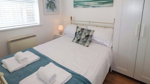 A bedroom with a bed, a bedside lamp, a painting, a radiator, and a window with blinds. - Sunrise Cottage (Chathill)