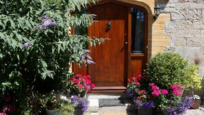 A wooden door with a knocker, flanked by potted plants with purple and pink flowers, and a stone wall. - Bell Cottage (Penzance)