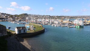 A coastal town with a harbor filled with boats, a small white building near the water, and a backdrop of houses and hills. - Bell Cottage (Penzance)