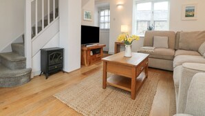 A living room with a staircase, a flat-screen TV, a wooden coffee table, a beige sofa, and a window with blinds. - Bell Cottage (Penzance)