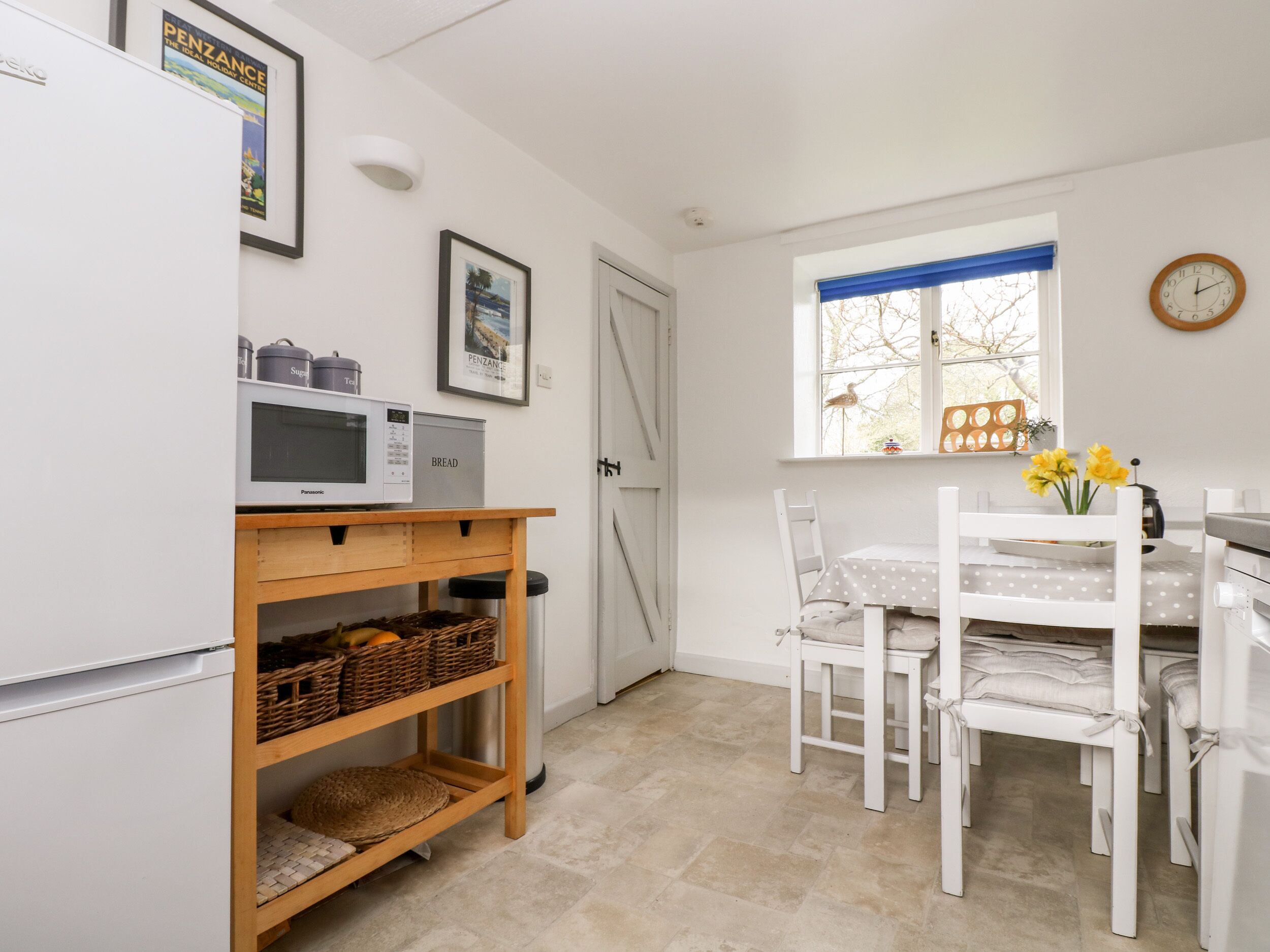 A compact kitchen with a microwave, bread box, and wicker baskets on a wooden shelf. A white table with chairs and a window with blue curtains are visible.