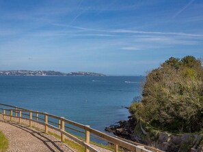 A coastal view with a wooden walkway leading to the sea, a rocky shoreline, and a distant town. - Bayview (Brixham)