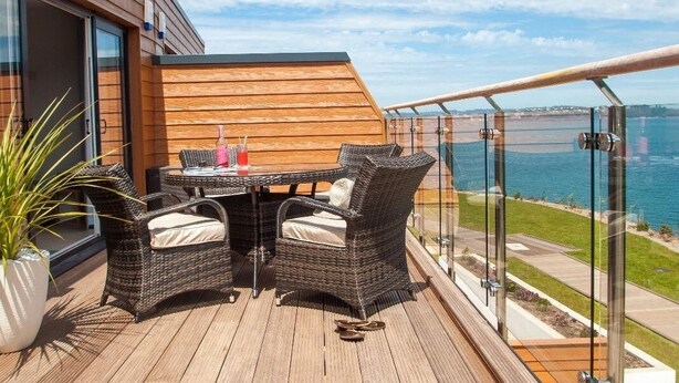 A wooden deck with wicker furniture and a glass railing overlooking a body of water.