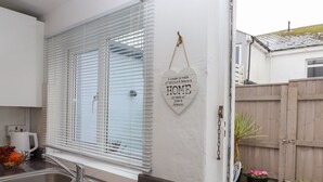 Une cuisine avec un évier en acier inoxydable, des armoires blanches et une fenêtre avec des stores. Un panneau décoratif accroché au mur porte l’inscription suivante : « Une maison est faite de briques et de poutres, un foyer est fait d’amour et de rêves. »