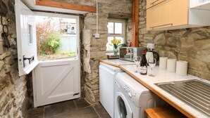 A compact kitchen with a stone wall, a washing machine, a microwave, and wooden cabinets. - Wayside Cottage (Consett)
