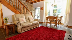 A cozy living room with a leather sofa, a red rug, a dining table with chairs, a wooden staircase, and a chandelier. - Wayside Cottage (Consett)