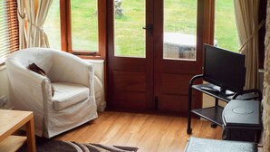 A room with a white armchair, a wooden table, a TV on a stand, and a view of a grassy area through a door. - The Granary (Ross-on-Wye)