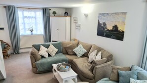 A cozy living room with a green and beige sectional sofa, a small white side table, and a wicker chair. A radiator is visible in the background, and a painting hangs on the wall. - The Coach House (Newport)